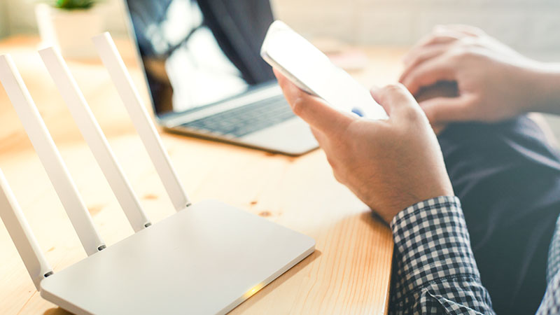 A man uses his phone next to a laptop and a wi-fi router.