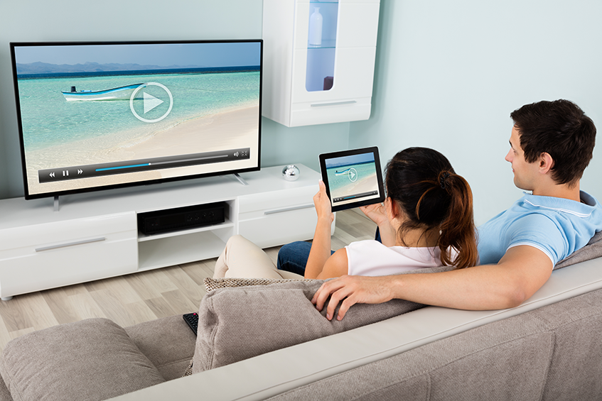 A couple sits on a couch while holding a tablet which is streaming to the television.