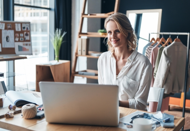 Woman sits behind her desk on her computer at her office while smiling.