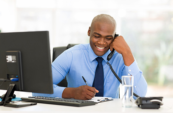 A man sits behind a desk with a computer writing down notes while talking on the phone.